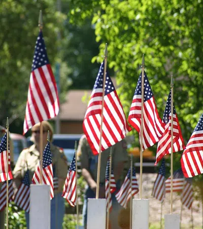 Multiple American flags on white posts with people blurred in the background on a sunny day