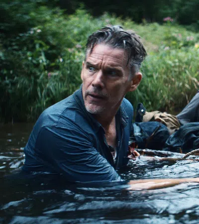 Two men cautiously wading through a river surrounded by dense greenery, holding onto a rope for support.