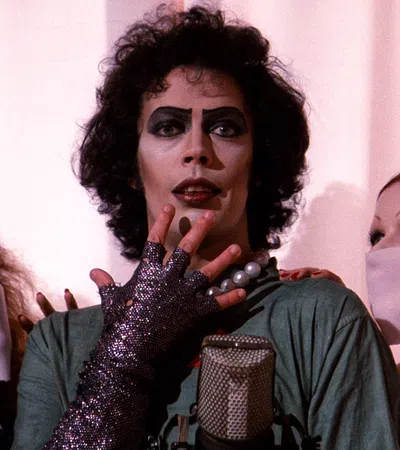 Actor in iconic glam rock makeup and costume with two masked fans on either side against a pale curtain.