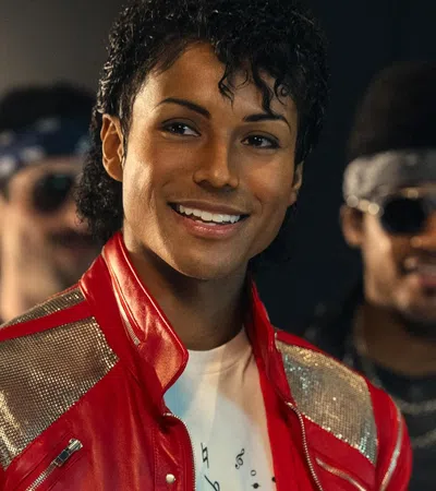 Smiling person wearing a red jacket with silver accents, with two others blurred in the background in dim lighting.