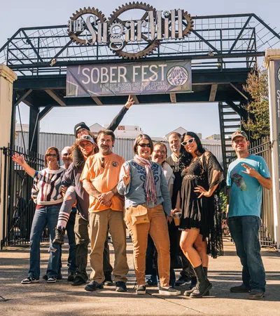 Group of people posing and smiling under Sober Fest banner at outdoor event entrance on sunny day