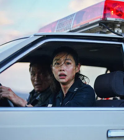 Female police officer and male colleague in a police car with red siren light, looking concerned through open window.