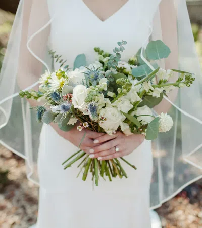 Bride in white dress holding bouquet of white and green flowers with veil outdoors on sunny day