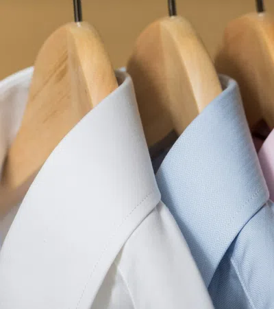 White, blue, and pink dress shirts neatly hanging on wooden hangers against a beige background