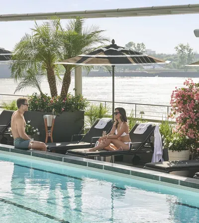 Couple relaxing and talking on lounge chairs by a pool with umbrellas and river view in the background