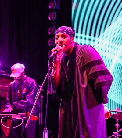 Hip-hop artist performing on stage with microphone and DJ in the background under vibrant purple and blue lights.