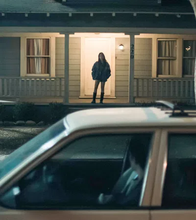 Person stands on lit porch of house at night with car parked in driveway in the foreground.