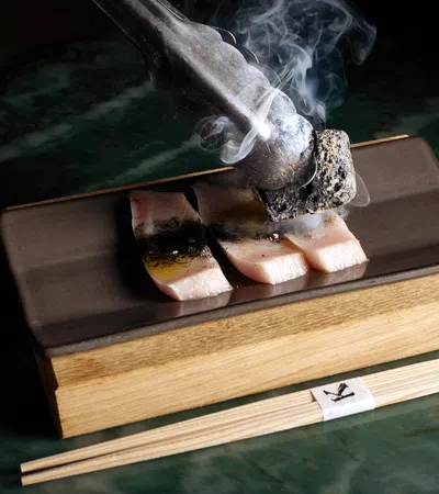 Close-up of sizzling fish slices being grilled with smoking charcoal on a rectangular plate with chopsticks.