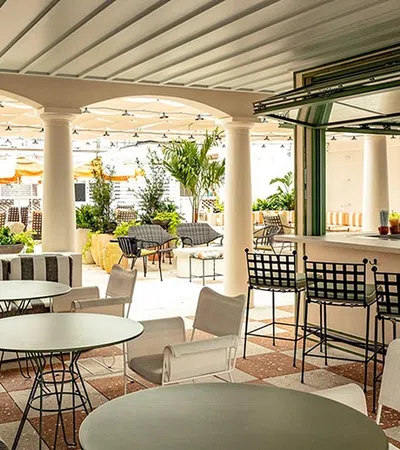 Bright outdoor patio with striped sofa, round tables, bar stools, and lush green plants under covered roof