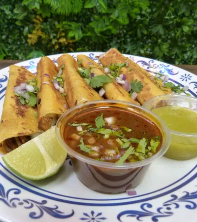 Plate of golden taquitos garnished with cilantro and onions, served with lime wedges and two dipping sauces.