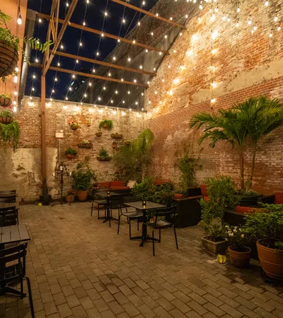 Cozy outdoor patio with string lights, plants, brick walls, and black metal tables and chairs at night