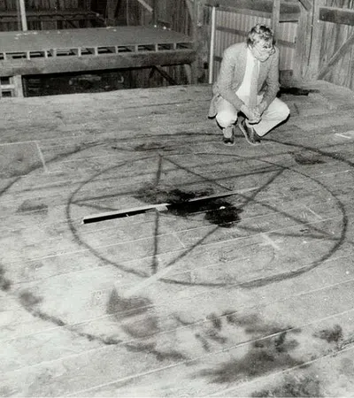 Black and white image of a man inspecting a large pentagram symbol drawn on a wooden floor inside a barn.