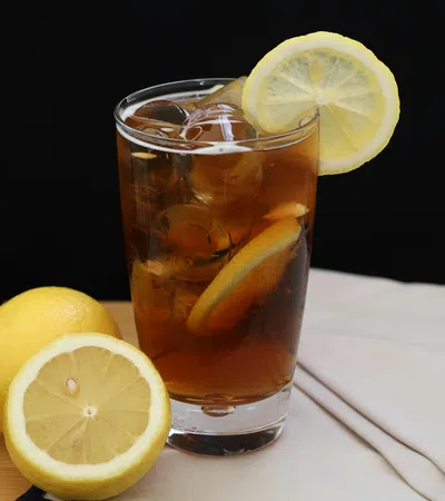 Glass of iced tea with lemon slices and whole lemons on wooden surface against black background.