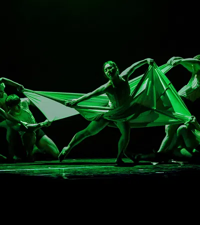 Performers in a dramatic green-lit dance holding and stretching a large fabric on stage in dynamic poses.