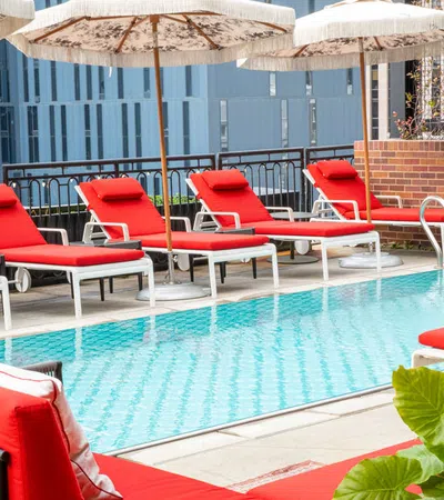 Outdoor pool area with red lounge chairs and umbrellas on a sunny day, surrounded by urban buildings.