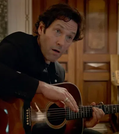 Man in black jacket playing acoustic guitar indoors with wooden doors and electric guitar in background