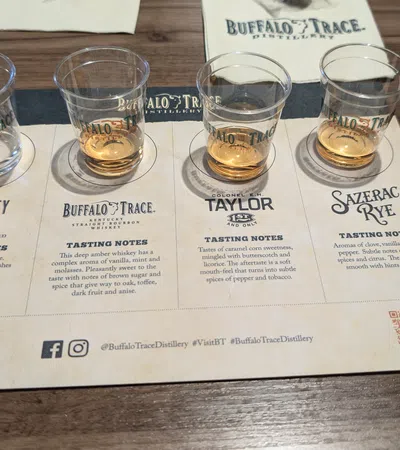 Four whisky tasting samples on a Buffalo Trace Distillery flight tasting mat with tasting notes and a cup on a wooden table.
