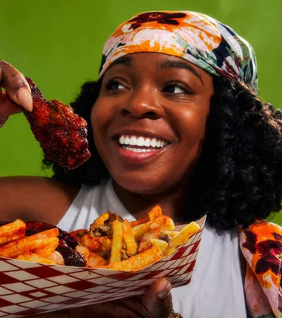 Smiling woman holding a saucy chicken wing and a tray of seasoned fries against a green background.