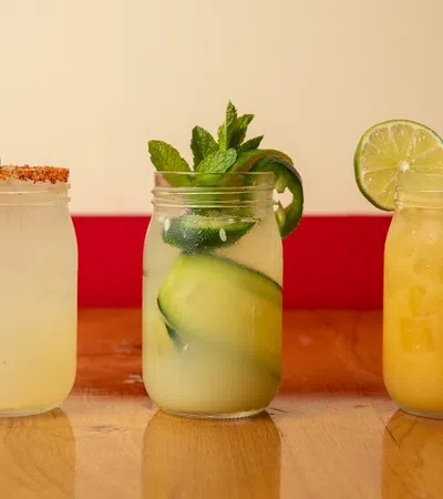 Three refreshing cocktails in mason jars with lime garnishes on a wooden table, featuring cucumber and mint.