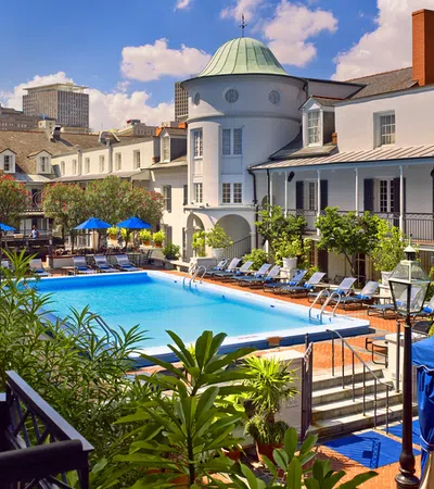 Outdoor pool area surrounded by lounge chairs, greenery, and historic white buildings under a blue sky.