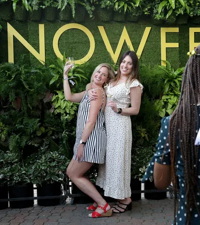 Two women pose and smile in front of a green plant wall with large yellow NOWEF letters while another person photographs them.