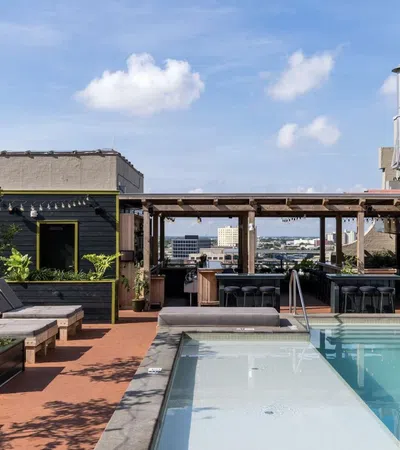 Rooftop pool with lounge chairs, greenery, and a covered bar area under a clear blue sky in an urban setting.