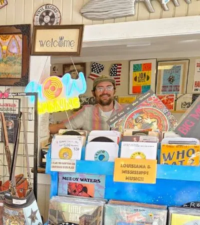 Smiling man behind record store counter with vinyl records, vintage posters, and neon open sign
