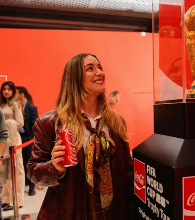 Woman holding Coca-Cola can looks at FIFA World Cup trophy in red-themed exhibit during trophy tour.