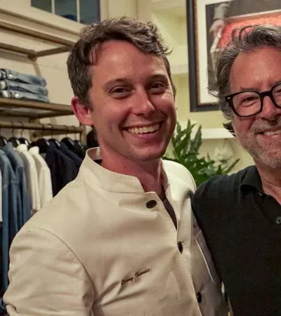 Two men smiling together indoors, one wearing a white chef jacket and the other in a black shirt with glasses.