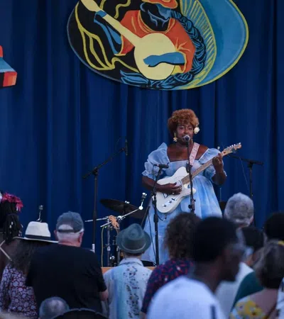 Singer with electric guitar performs on stage in front of crowd at outdoor music festival with blue backdrop.