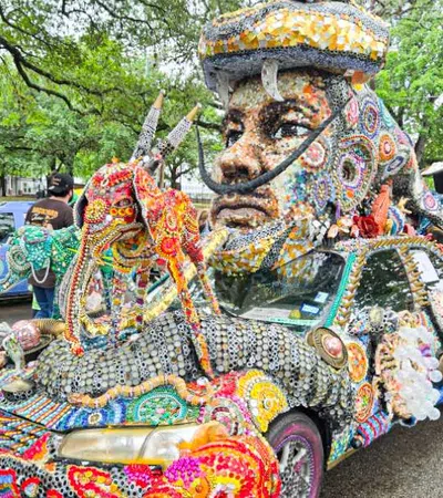 Colorful mosaic-covered car with a large head sculpture on top at an outdoor event with people and trees.
