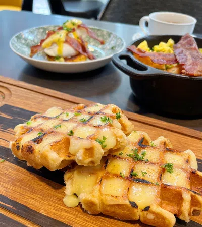 Two grilled cheese waffles garnished with herbs on a wooden board with breakfast dishes in the background.