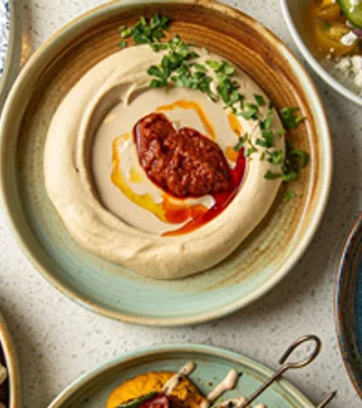 Plates of Middle Eastern dishes including hummus with chili oil, pita bread, salad, and roasted vegetables on a table.