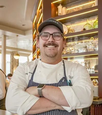 Smiling chef with glasses and cap stands with arms crossed in a restaurant kitchen with staff working in the background