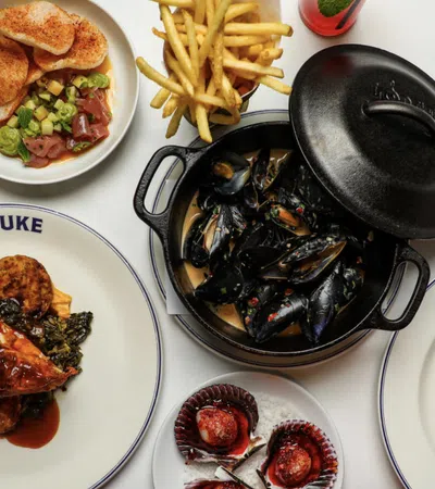 Variety of gourmet dishes including mussels, grilled chicken, fries, and desserts on white table with LUKE plate.