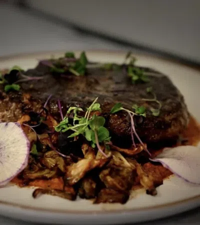 Grilled steak served with sautéed mushrooms, microgreens, and thinly sliced radish on a white plate.