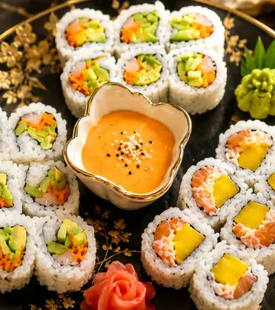 Assorted sushi rolls with avocado, salmon, mango, and dipping sauce on a decorative black plate.