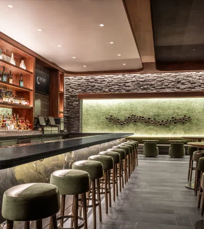 Modern stylish bar interior with green velvet stools, marble counter, illuminated shelf, and textured stone wall.
