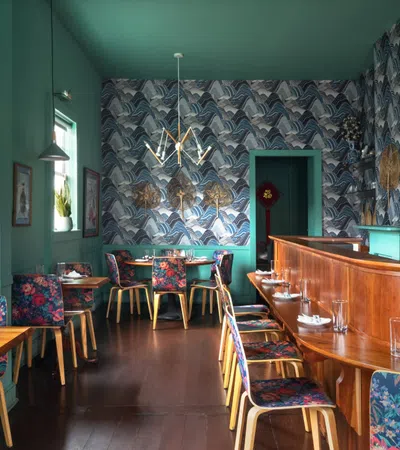 Cozy restaurant interior with teal walls, floral chairs, wood bar, and patterned wallpaper under modern lighting.