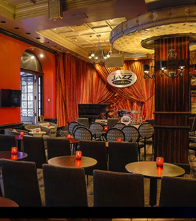 Cozy jazz club interior with stage, drum set, piano, round tables, candles, and warm red and orange decor.