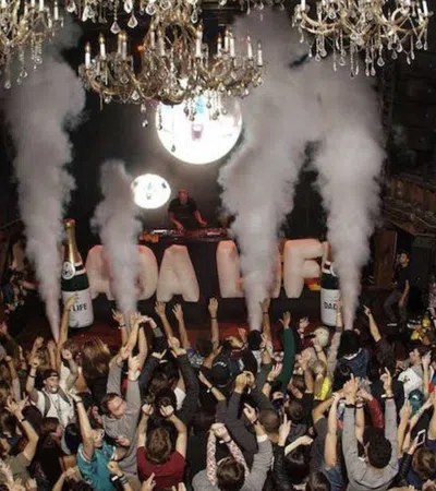 Crowded nightclub with people celebrating under chandeliers and large smoke effects from bottles on the dance floor.