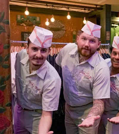 Four men wearing retro diner uniforms and hats posing with outstretched hands in a cozy restaurant setting.