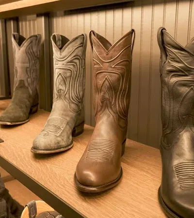 Row of six cowboy boots in various shades displayed on a wooden shelf against a paneled wall.