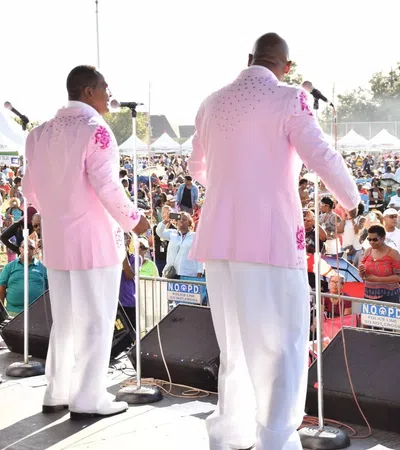 Five male singers in pink jackets performing on outdoor stage before large audience at daytime event.