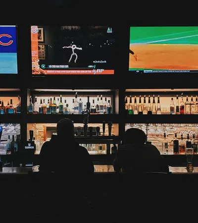 Silhouetted patrons watch multiple sports games on TVs behind a dimly lit bar with liquor bottles on shelves.