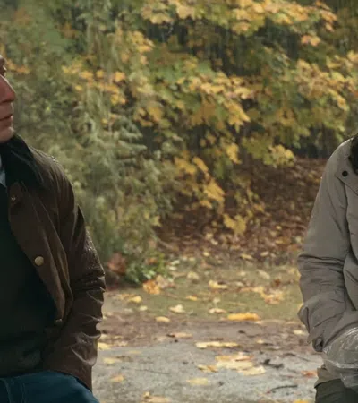 Man and woman dressed in autumn clothing sit outdoors surrounded by fall foliage during a conversation.