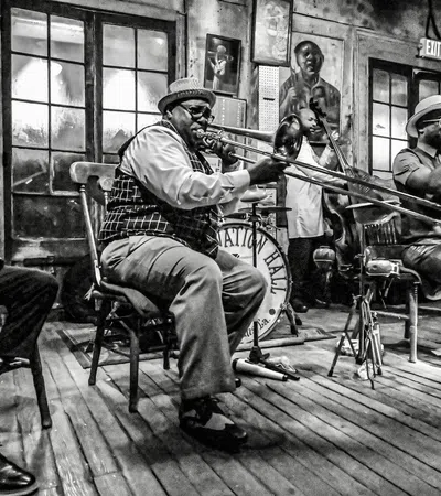 Black and white photo of jazz musicians playing saxophone, trombone, and trumpet on wooden floor stage indoors.