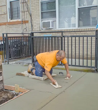 Construction worker smoothing fresh concrete on a sidewalk while another man watches at a residential building.