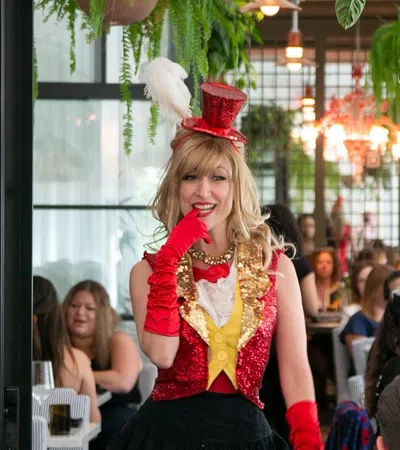 Woman in red sequined vest and hat with feather poses playfully at crowded restaurant event.