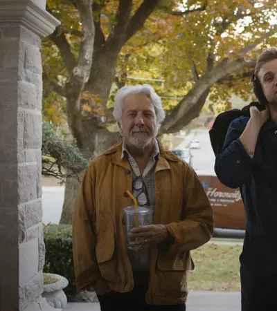 Older man with drink and younger man with headphones and backpack standing on porch in autumn setting